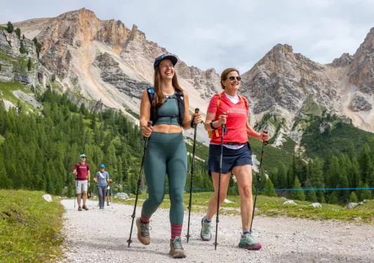 Two women smiling while walking on a rocky trail, with tall mountains in the distance