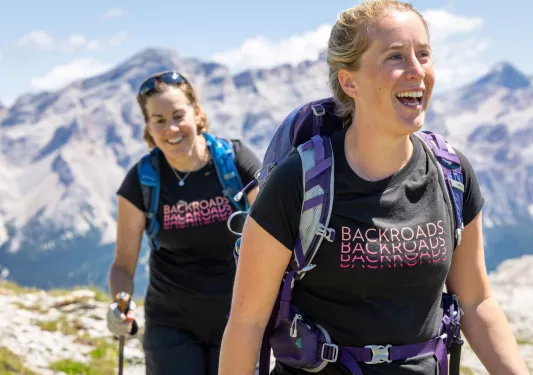 Two women smiling, wearing backpacks while hiking on a mountain