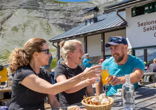 Two women and one man smiling while raising their glasses of alcohol, sitting outside