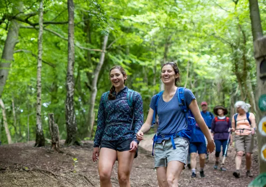 Two women walking along a dirt trail with tall trees in the background