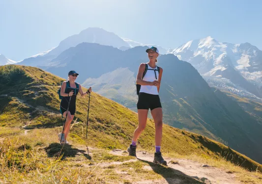 Two women smiling, while hiking on a dirt trail on top of a mountain