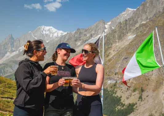 Three women smiling next to an Italian flag, with large mountains in the background