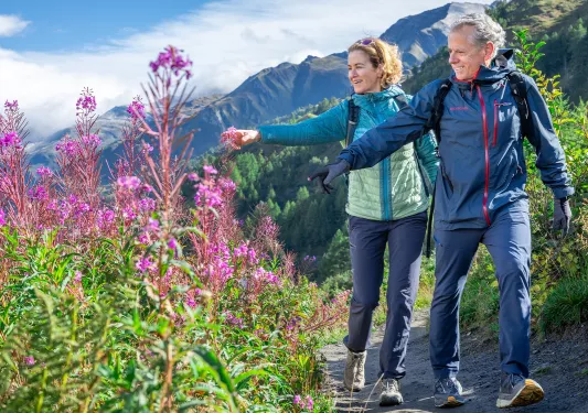 Man and woman walking on a dirt trail while touching pink flowers