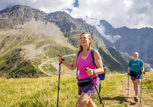 Two women walking on a dirt trail, with large mountains in the background