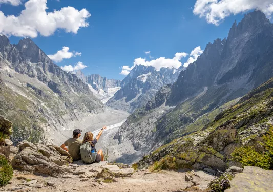 Man and woman sitting on a cliff, pointing out to large mountains in the distance