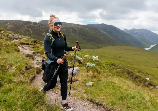 Woman with hiking poles, walking on a dirt trail with a grass valley in the distance