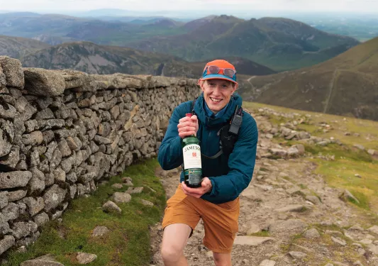 Man holding up a bottle of Jameson, walking next to a large stone wall