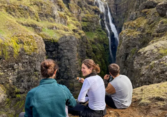 Three people sitting on a cliff, with mountains and waterfalls in the distance