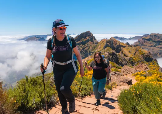 Two women smiling, while ascending a stone path on a mountain with hiking poles