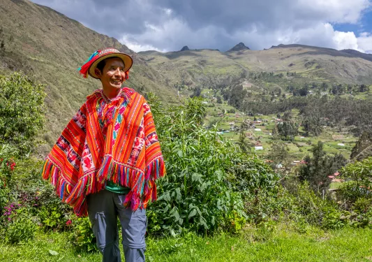 Man wearing traditional attire, in a grass field with hills and houses in the background
