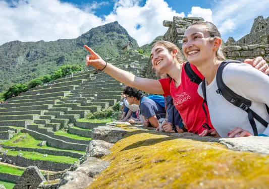 Two women smiling and pointing towards the sky, with large stone ruins in the background