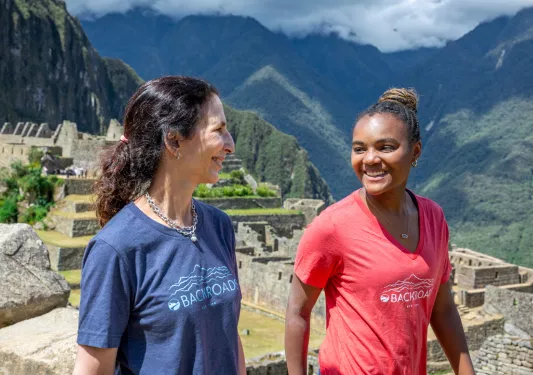 Two women smiling and looking at each other, while walking on a trail with mountains in the background