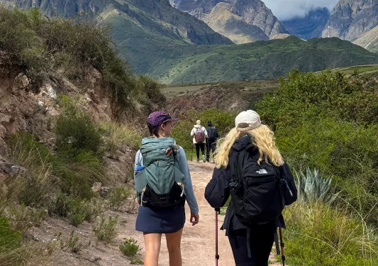 Two women walking on a dirt trail, with large hills and mountains in the distance
