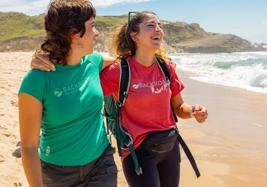 Two women smiling with their arms around each other, walking on the beach