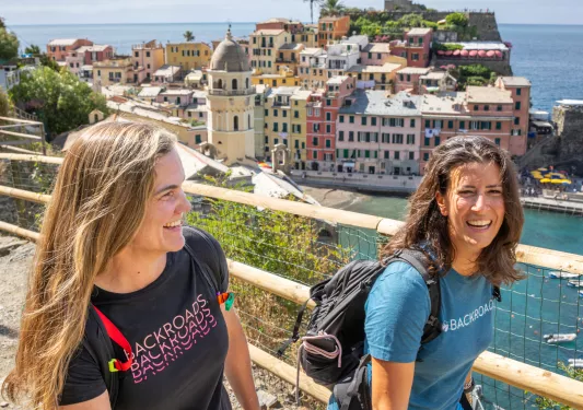 Two women smiling while walking along a dirt path with houses in the background