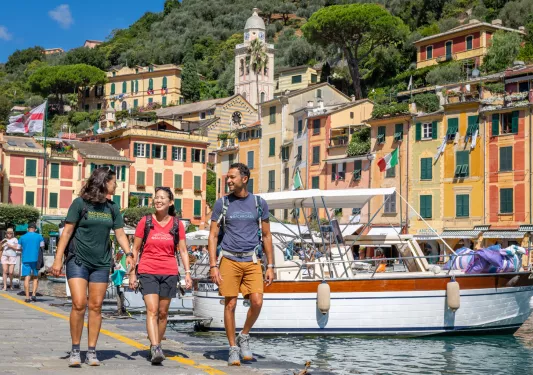 Two women and one man walking along a boat dock, with orange buildings in the background