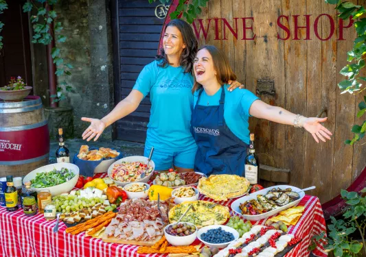 Two women smiling with their arms open, behind a table full of food