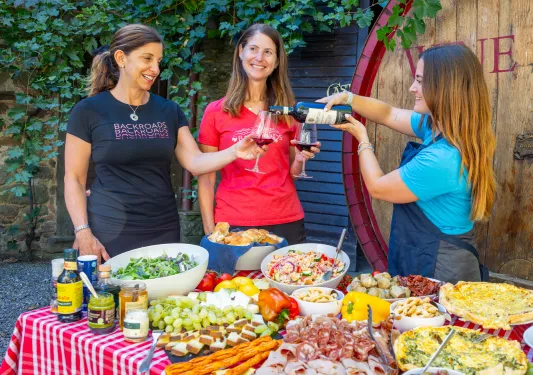 Three women behind a table full of food, pouring wine into glasses
