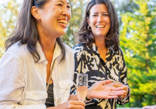 Two women smiling while holding glasses of drinks,, with tall trees in the background