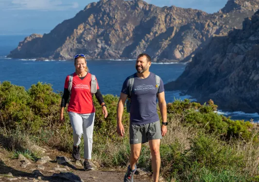 Man and woman smiling while walking on a dirt and stone path and the ocean in the background