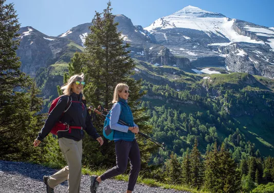 Two women hiking along a rocky trail, with snow-capped mountains in the background