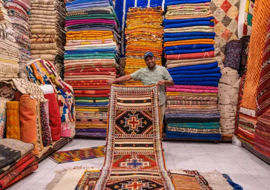 Man holding up a large rug in a room full of stacked rugs