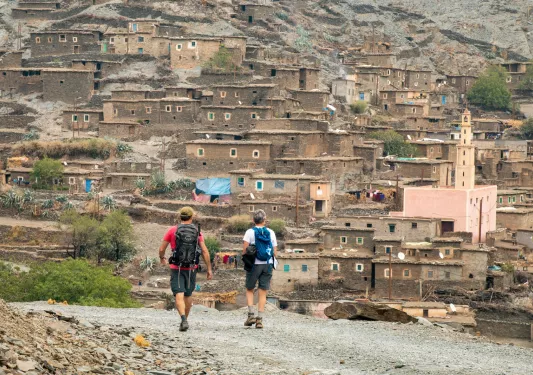 Two people walking on a dirt trail towards dirt and stone houses