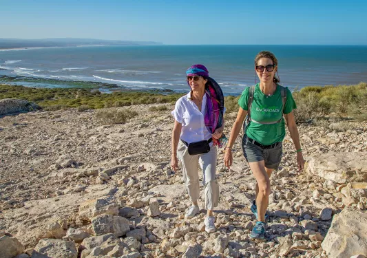 Two women smiling while walking on a gravel trail with the beach in the background
