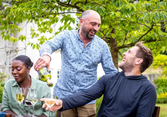 Man standing over a man sitting down, pouring him a glass of wine