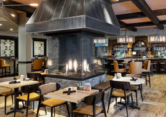 Restaurant dining area with wooden chairs and tables, with a large, enclosed firepit in the center