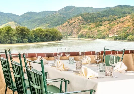Dining table with green chairs, next to a river surrounded by trees and hills