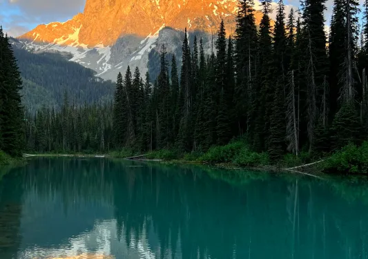 Tall mountains hit by the sunset, with a lake surrounded by tall trees