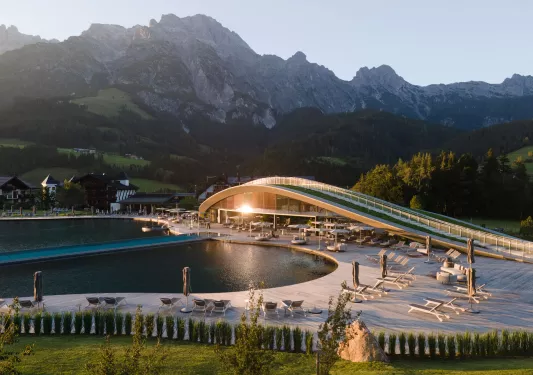 Large building with a large pool in front, and tall mountains in the background