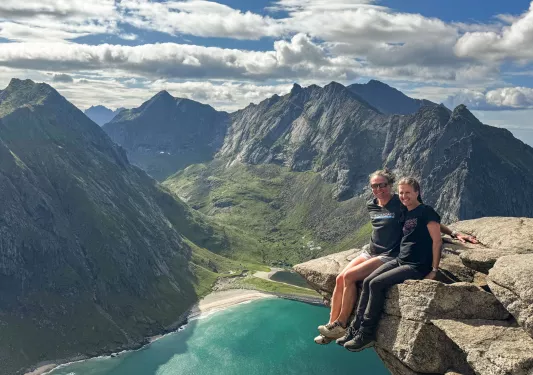 Two women sitting on the edge of a rock with a lake and mountains in the distance