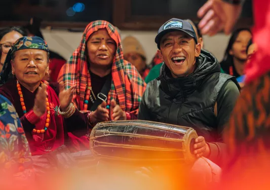 Man smiling while playing a hand drum, with a group of women clapping behind
