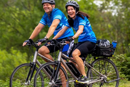 Two guests biking on forest road, smiling at camera.