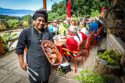 Local chef with meat platter, guests at table behind them.