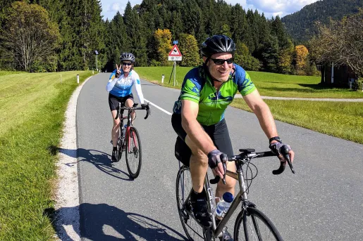 Two bikers riding down a hilly, tree lined street.