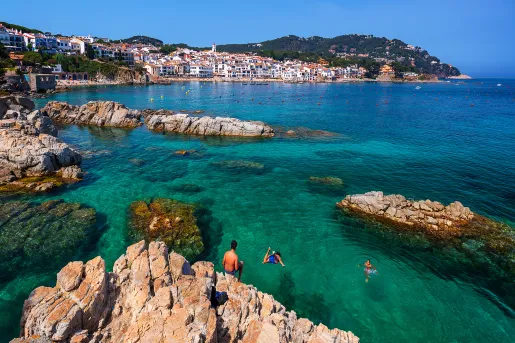 Guests jumping off craggy cliffs into blue water, Costa Brava in distance.