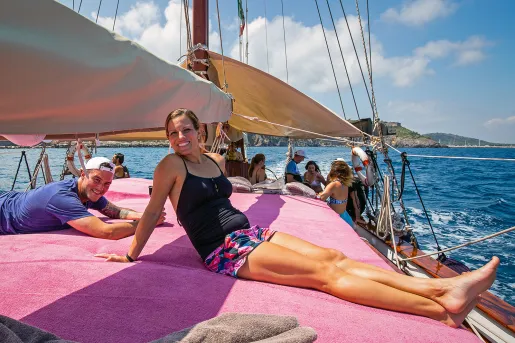 Group of guests relaxing on sailboat, coastal town in background.