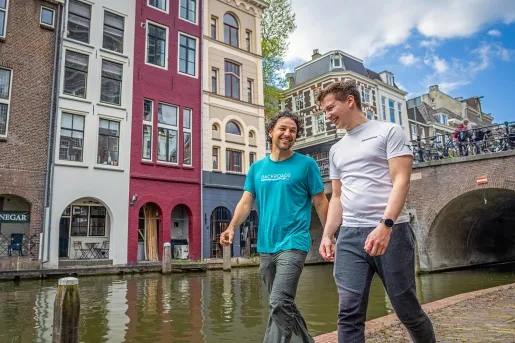 Two guests walking next to a river in town, colorful storefronts behind them.