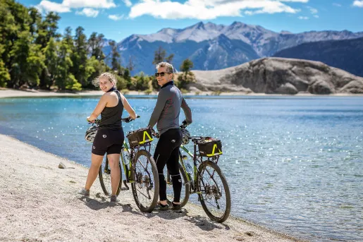 Two guests with bikes, walking along beach, blue water, forest behind them.