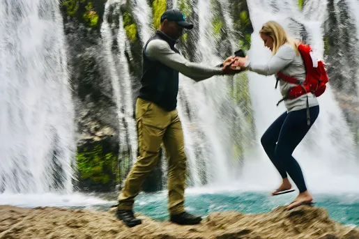 Two guests walking over rocks, waterfall behind them.