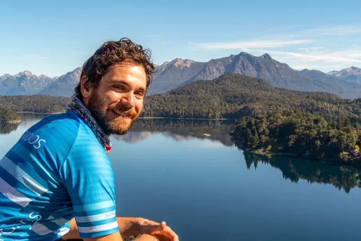 Guest posing next to still lake, forest, mountain behind them.