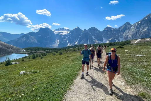 Group of guests walking next to small river, grassy meadow, mountains in background. 