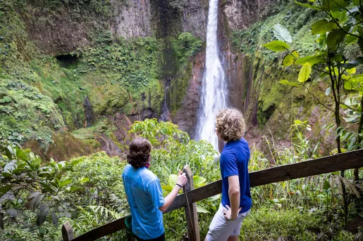 Two guests on forest path, looking down at forest, waterfall.