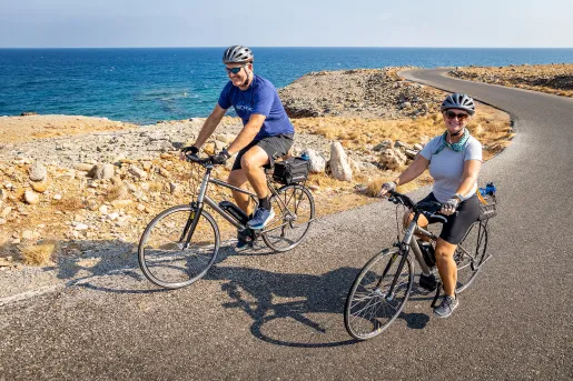 Two guests biking alongside golden coastline, smiling at camera.