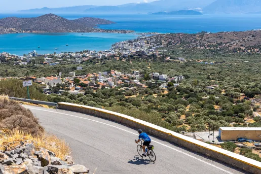Guest cycling on road, arid coastal town in distance, ocean behind it.