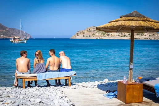 Four guests on rocky beach sitting on bench, overlooking ocean, one looking back at camera.