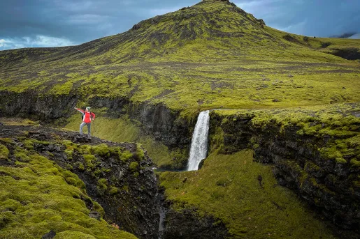 Guest in hiking gear amidst green mountainside, peak, waterfall behind her.
